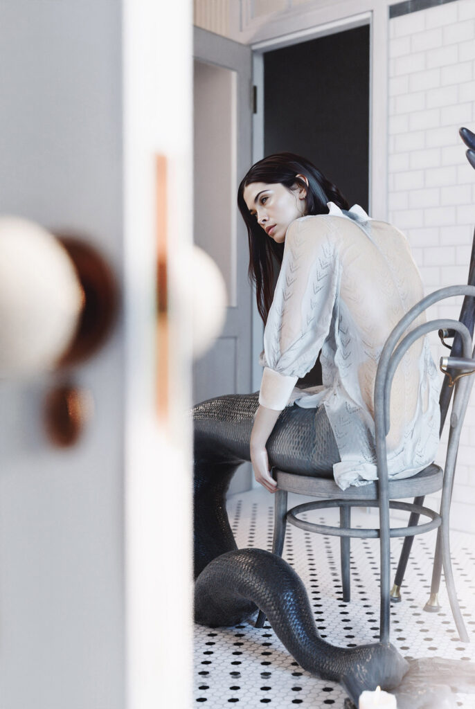 A woman with a mermaid tail sits on a chair in a room with white tiles and a doorway.
